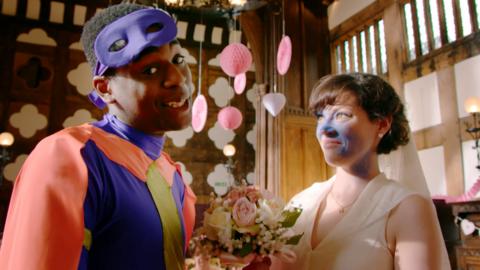 A bride with a blue face and a groom wearing a superhero outfit stand smiling at the altar.