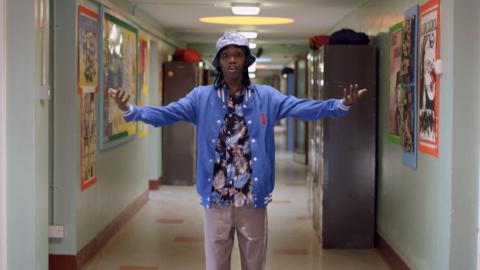 Boy in a blue jacket and hat stands with his arms raised in a school corridor.