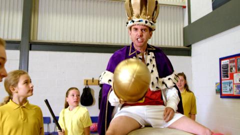 A man with a crown, sceptre and robe sits across a pommel horse.