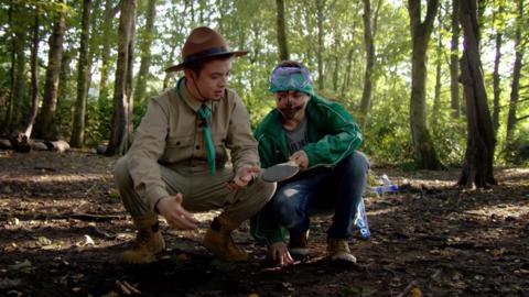 Two boys crouch together in a forest, staring at a table tennis paddle.