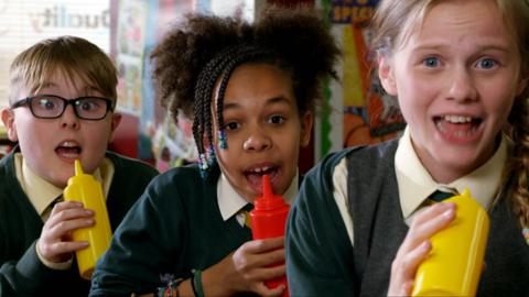 Three school kids sing into ketchup and mustard bottles.