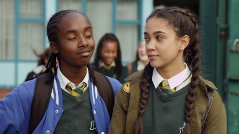 Boy smiles slyly at girl who looks suspicious outside school gates.