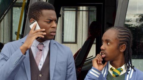 A man and a boy look at each other in surprise while standing outside a white bus on mobile phones.