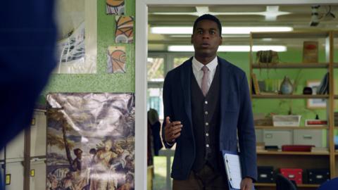 A man in a smart suit stands in a teacher's staff room with a clipboard.