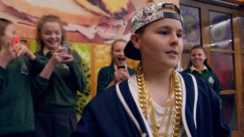 A boy with a backwards cap and gold chains looks confident as girls behind him point and stare.