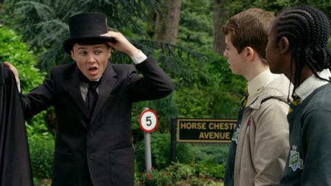 A boy with a top hat, black suit and cape stands in front of two boys looking on.