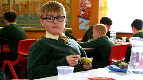 A boy with glasses holding a banana smiles with his eyes raised.