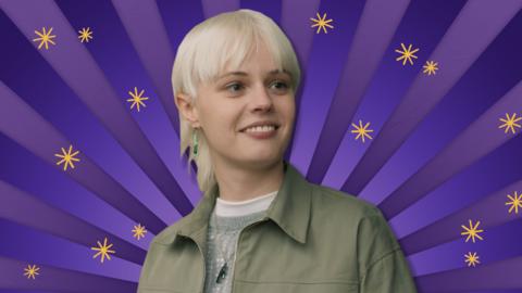 A blonde girl wearing a green jacket standing in front of a purple background whilst smiling