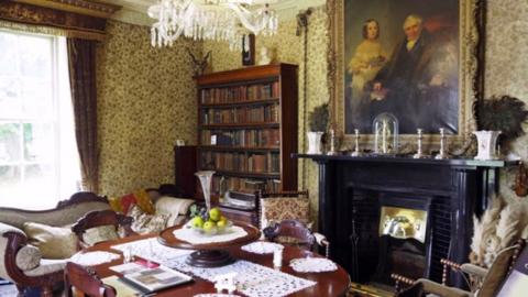 The interior of a study with a large portrait of an old man on the wall and books on a bookcase.