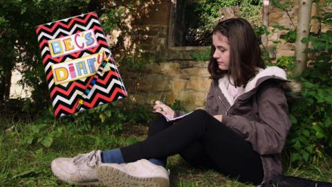 A girl writes in her diary sat in a garden, Bec from The Dumping Ground.