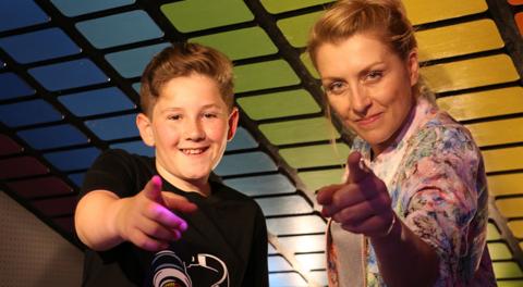 A boy and a woman point to the camera in front of a DJ charts graphic.