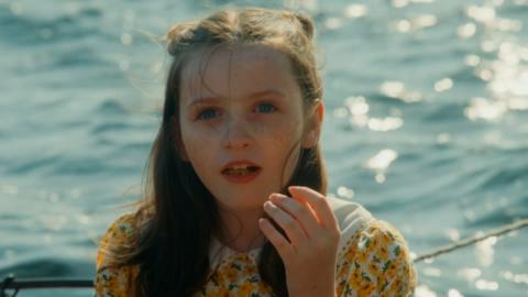 A young white girl with long brown hair is wearing a yellow flower pattern dress and standing next to the ocean