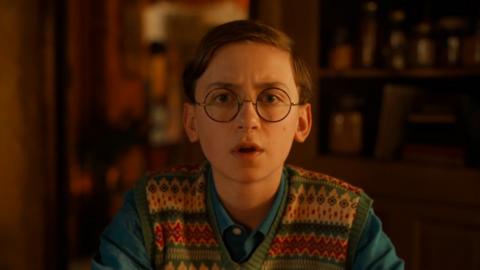 A young white boy with short brown hair is wearing black circular glasses and a cardigan and is standing in a library