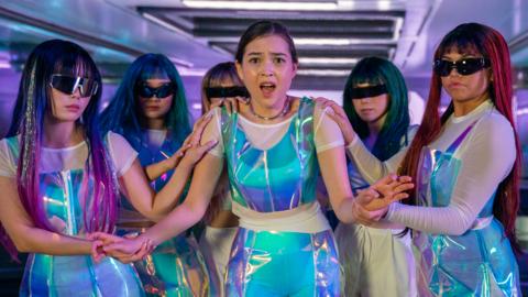 Hannah, a teen girl is dressed in blue shiny outfits, looking confused whilst five females dressed the same but wearing sunglasses surround her.