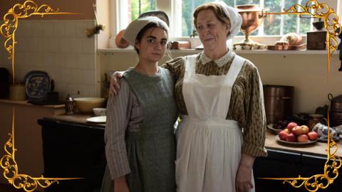 A elderly lady in cook's uniform had her arm around a young girl, also in serving clothes, as they stand in a kitchen, set in a gold frame.