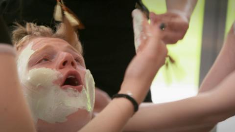 A boy cries out as girls attack him with a face mask and make up.