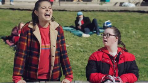 Marnie from Mustangs FC looks on from the side of a football game, she's cheering support with a friend.