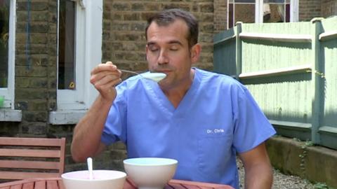 Dr Chris sitting at a table and holding a spoonful of yoghurt to his mouth.