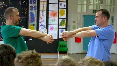 Dr Xand and Dr Chris both hold their arms out in front of them. Their arms are outstretched and crossed over with fingers interlocked. They are standing in a school gym.