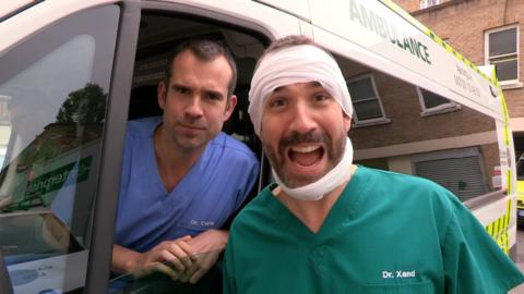 Two doctors by an ambulance, one has bandages around his head and looks excited.