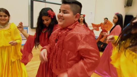 Boys and girls in brightly coloured clothing dance together in a hall.