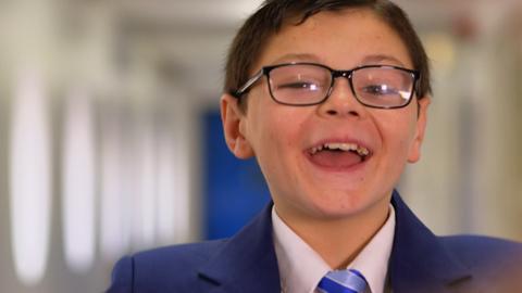 A boy with glasses laughs while standing in a long school corridor.