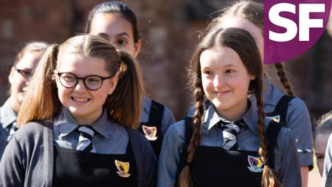 Mildred and Maud, two school girls are standing in their uniforms, smiling.