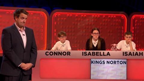 Four students sit behind a panel, whilst their teacher stands next to them.