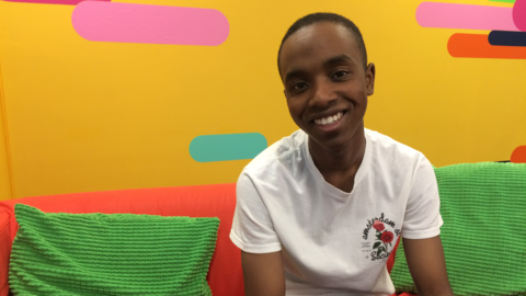 Akai Osei (Nero Johnson) sits and smiles at the camera against a bright yellow background.