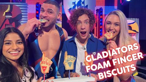 Shini and Joel from Blue Peter are stood in between Steel and Diamond from Gladiators. Shini is holding some red and blue decorated foam finger biscuits. Diamond and Steel are pretending to eat them.