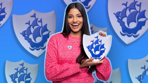 Blue Peter presenter Shini is wearing a pink jumper and holding a large blue Blue Peter badge. Behind her are multiple enlarged blue Blue Peter badges.