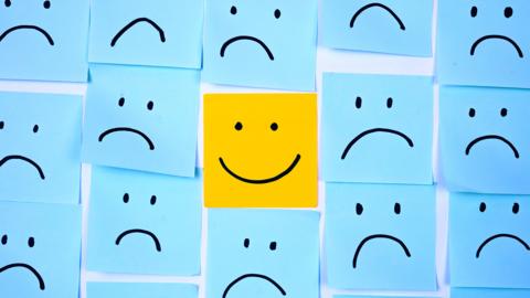 A wall covered in blue post it notes with sad faces on them. In the centre, sits a yellow post-it note with a smiley face.