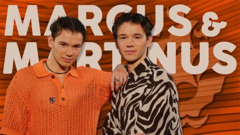 Marcus and Martinus, two young men with brown curly hair, they are brothers. On the left, he is wearing an orange shirt and on the right he is wearing a black and white zebra stripe shirt.