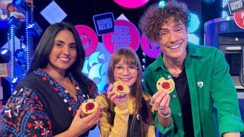 Three people are each holding a biscuit with a jam circle in the middle. From left to right: Rav, an Asian woman. Tilly, a young white girl. And Joel, a tall white male.