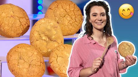 Great British Bake Off contestant Tasha Stone is smiling in a pink shirt whilst holding a wooden spoon in one hand and a white chocolate chip cookie in the other.