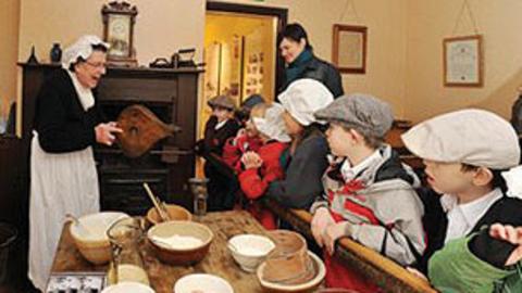 Children in hats watch a woman dressed in nineteenth century clothes use bellows.