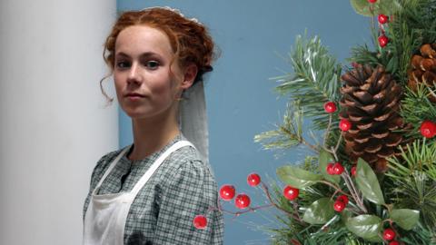 A girl (Hetty feather) smiles next to some Christmas holly.