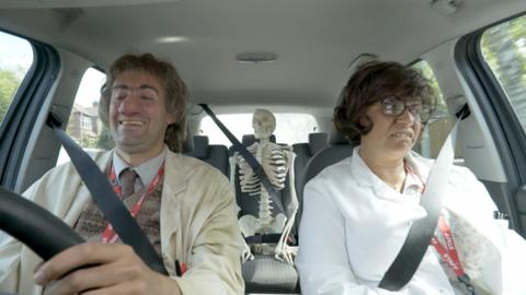 A lady and a man driving in a car. The lady looks annoyed.