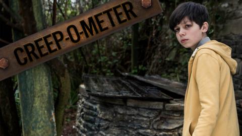 A boy in a yellow hoodie stands next to an old well looking tense.