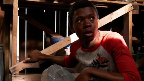 A boy crouching beside a table in a shed, hiding in the shadows folding a dustbin lid as a shield.