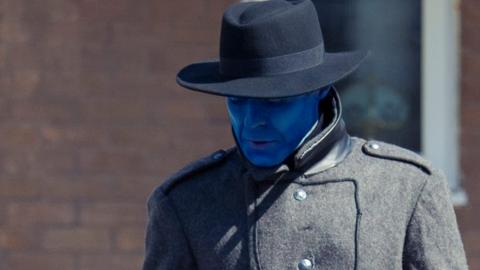 A man with blue skin wearing a black coat and hat looks down at the ground.