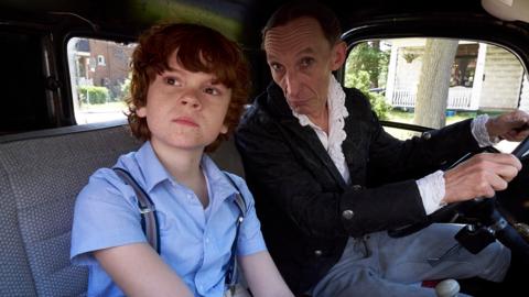 A boy with ginger hair sits in a car with a man while they both look out of the window.