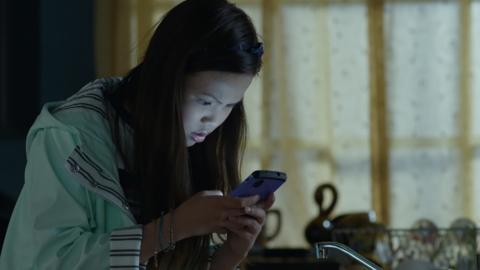 A girl with long dark hair is leaning over a mobile phone screen that lights up her worried face.