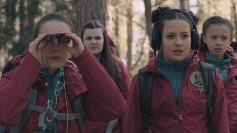 Two young girls in red jackets, one looking through binoculars and one wearing headphones.