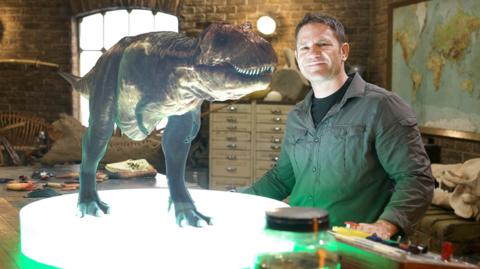 A man sat next a table that's lit up with a hologram of a T-Rex.