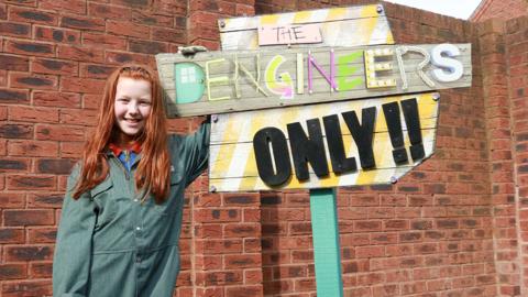 A young girl wearing overalls stands next to a sign saying Dengineers only.