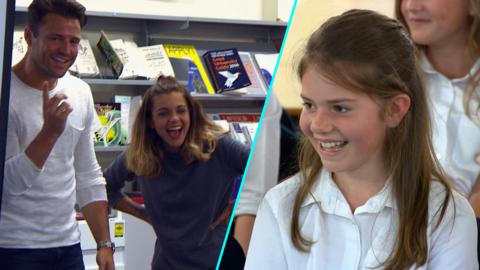 Mark and Lauren stand next to a projector screen at a school and surprise a young girl.