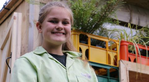 A girl smiles stood in front of a den built out of recycled materials.