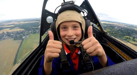 A boy grins, holding both thumbs up, while sitting in the cockpit of a flying plane.