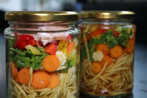 Two jars full of noodles and vegetables.